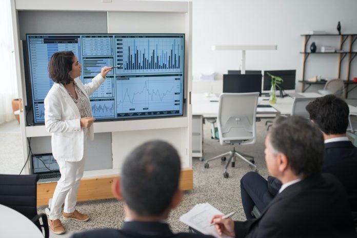 lady presenting at a monitor full of graphs and charts to small audience