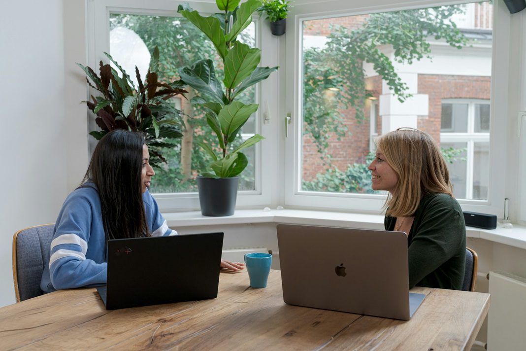 two women sitting at a conference table with laptops chatting